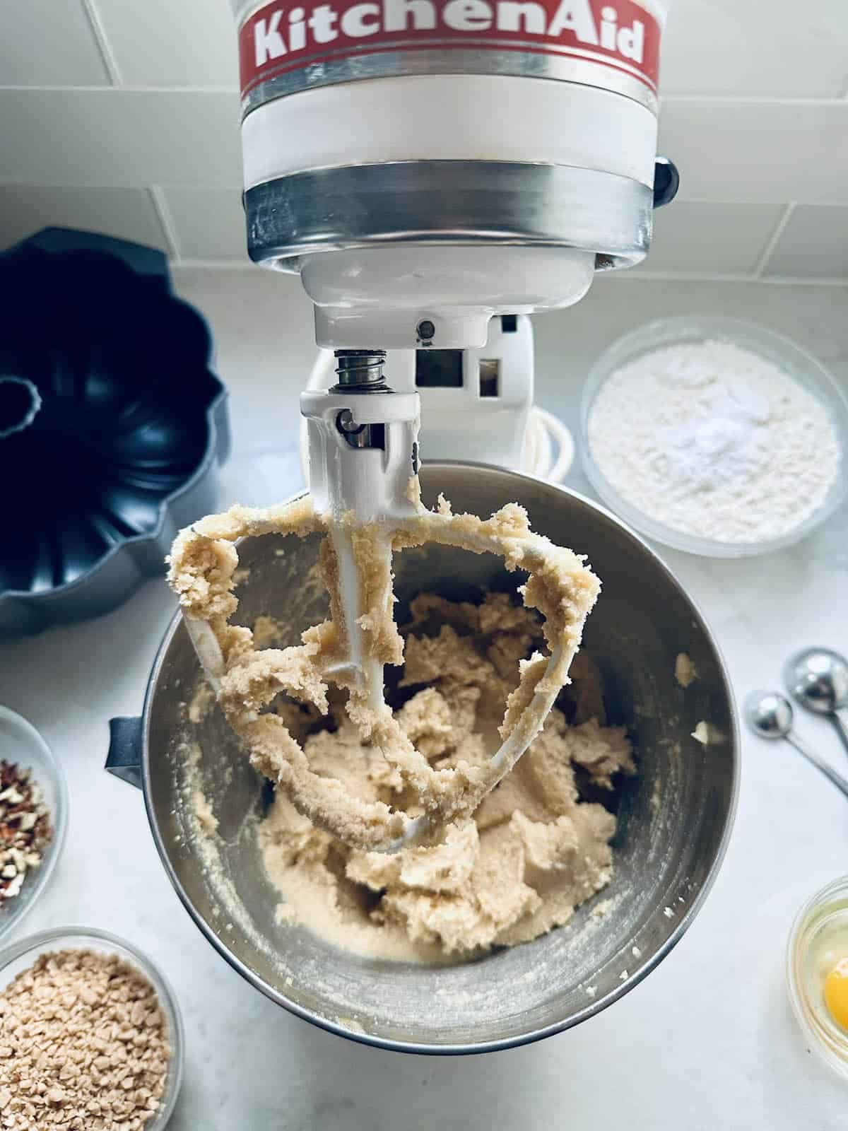 A stand mixer with cookie dough on the paddle attachment, surrounded by baking ingredients in bowls, including flour, nuts, and eggs, on a white kitchen countertop.