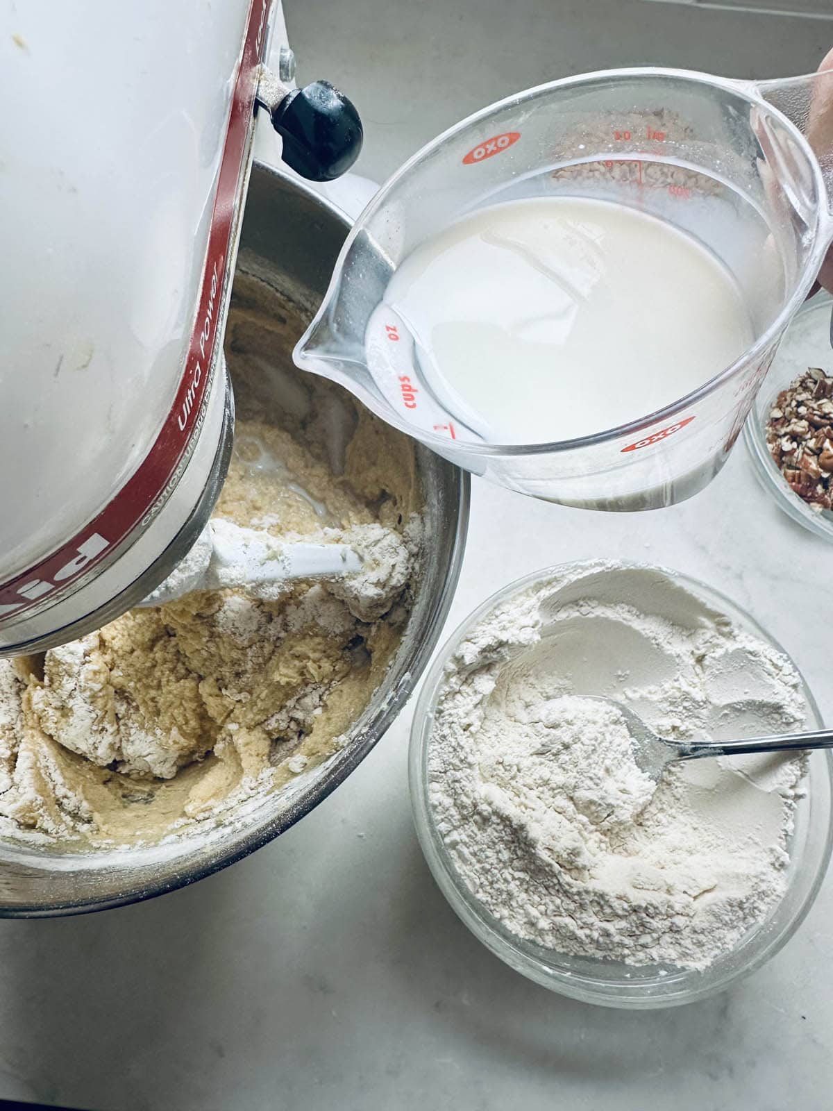 A person holding a measuring cup of milk above a stand mixer bowl with batter, next to a glass bowl of flour with a spoon and a small bowl of chopped nuts on a white countertop.