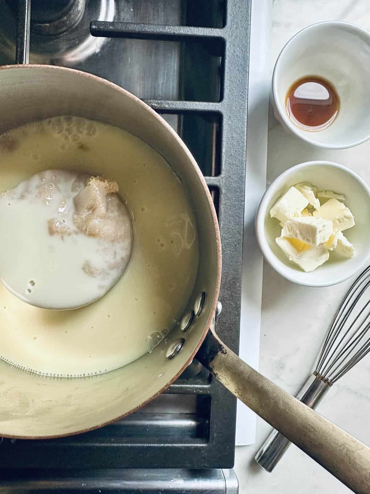 A saucepan on a stove with milk and brown sugar inside, next to small bowls containing butter and vanilla extract, and a whisk on a marble countertop.