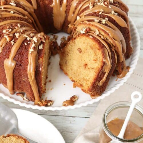 A bundt cake with caramel glaze and chopped pecans on top sits on a white plate. A slice has been cut and placed nearby, with a jar of caramel sauce and a spoon next to the cake.
