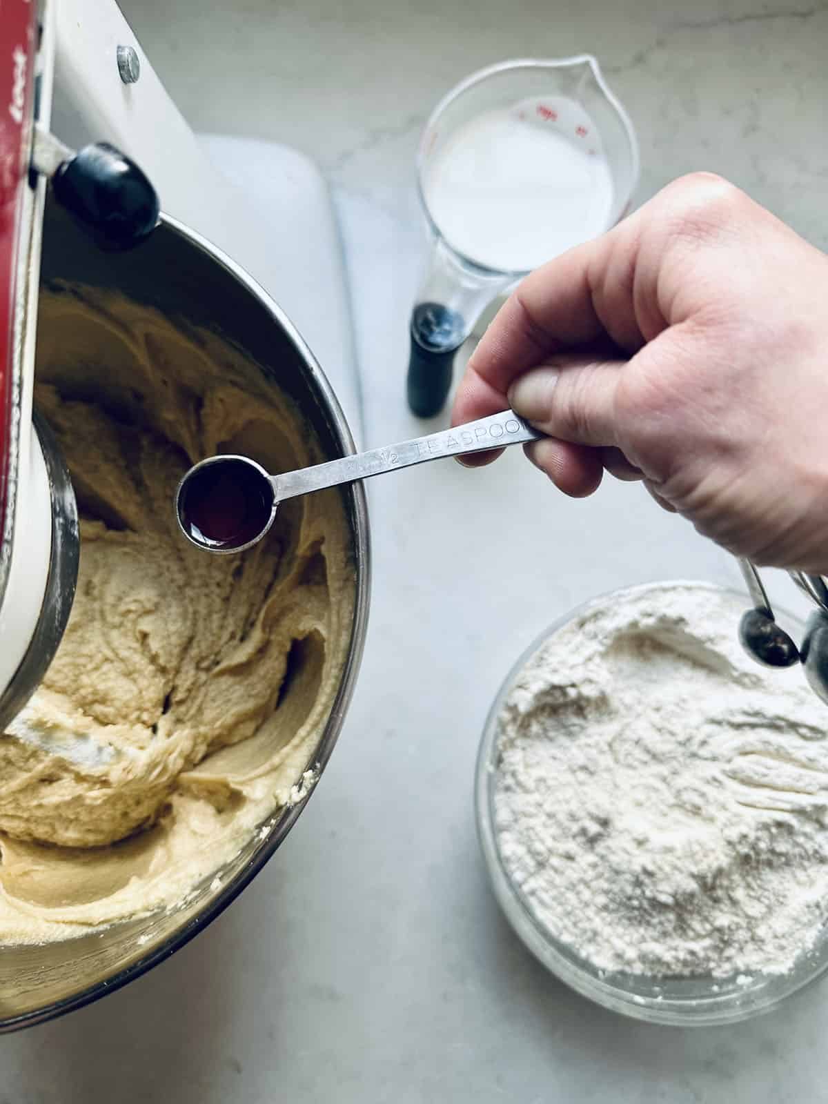 A hand holds a teaspoon of vanilla extract over a mixing bowl with batter. Nearby, a glass bowl of flour and a measuring cup of milk sit on a light countertop.
