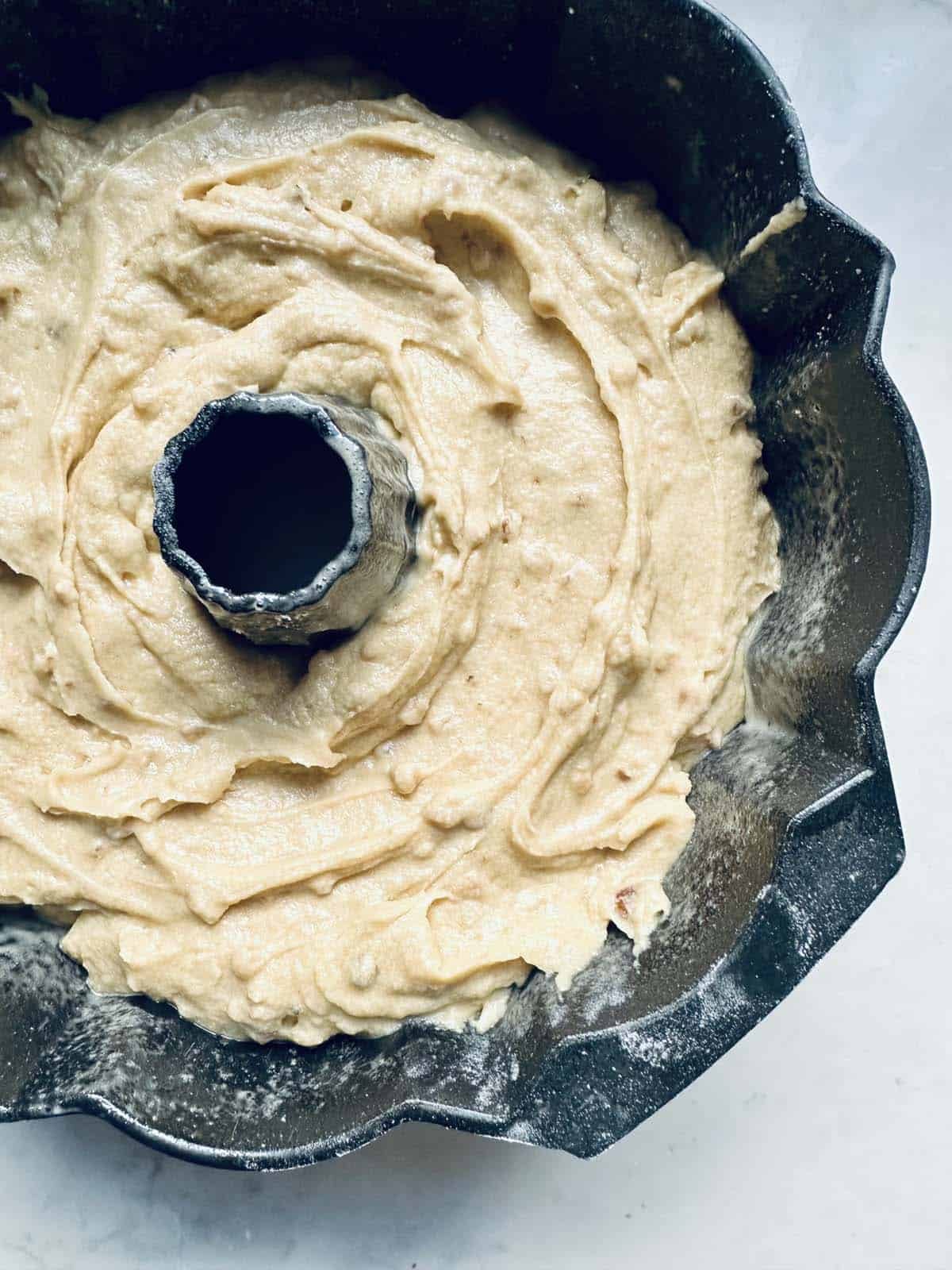 Cake batter spread evenly in a fluted bundt pan, ready to be baked. The pan sits on a light-colored surface, and the batter is creamy with a slightly textured appearance.