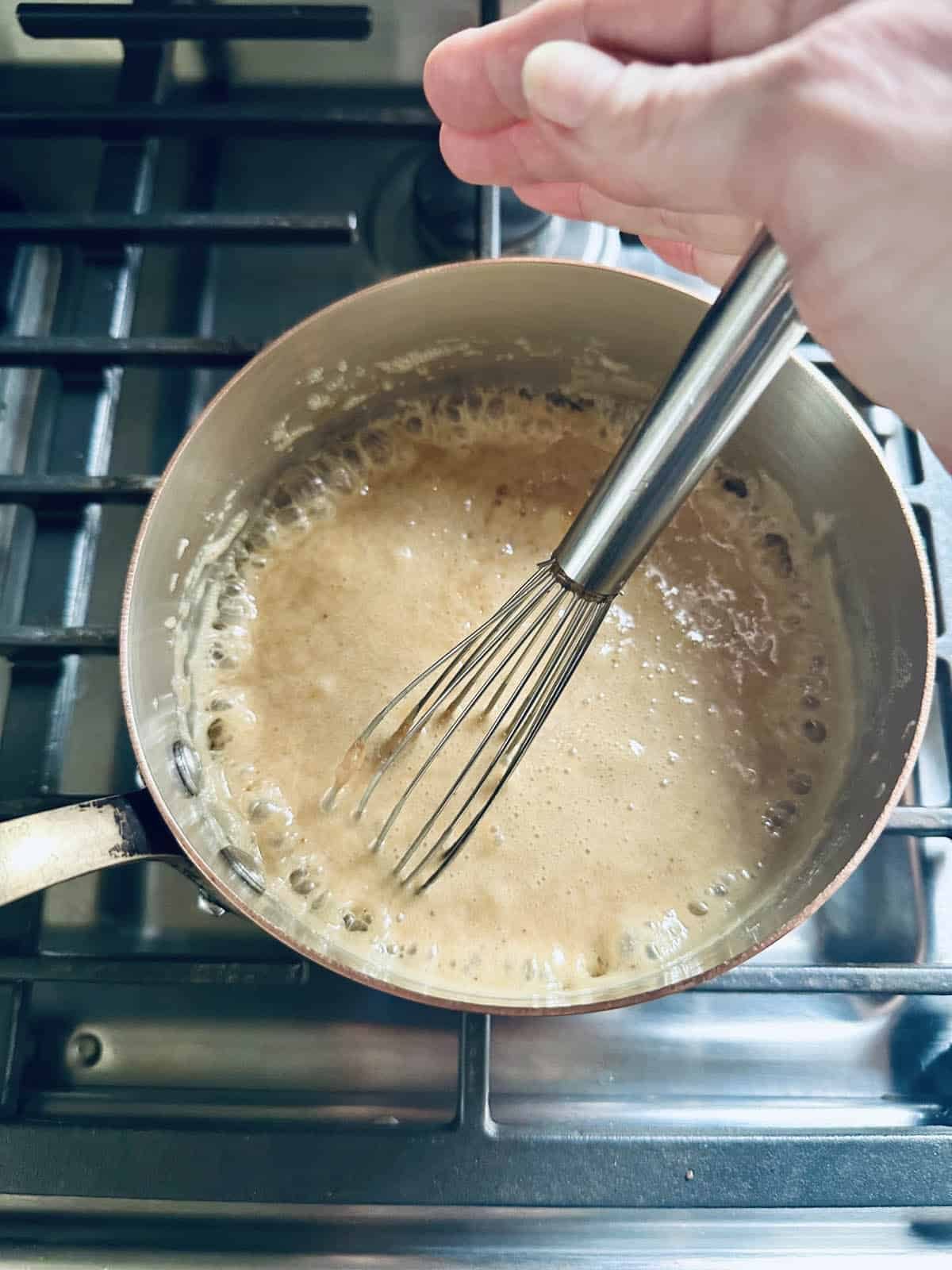 A hand sprinkles seasoning into a bubbling mixture in a saucepan on a stove, while stirring with a metal whisk.