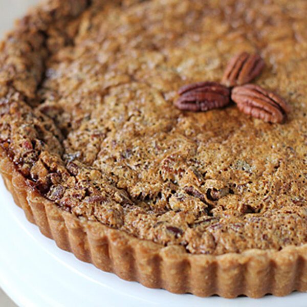 A close-up of a pecan pie with a golden-brown crust, topped with three whole pecans in the center, displayed on a white cake stand.