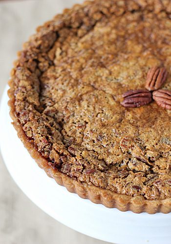 A close-up of a pecan pie with a golden, textured crust, topped with a small cluster of whole pecans in the center, displayed on a white cake stand.