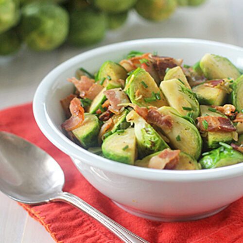 A white bowl filled with sliced Brussels sprouts and pieces of crispy bacon sits on a red cloth napkin, next to a silver spoon. A bundle of fresh Brussels sprouts is blurred in the background.