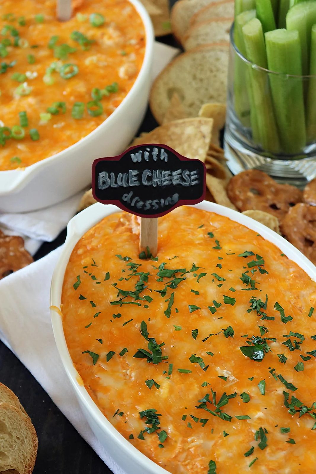 Oval dish of baked buffalo chicken dip, topped with chopped herbs, with a small sign reading with blue cheese dressing. Surrounding the dish are sliced bread, pretzels, tortilla chips, and a glass of celery sticks.