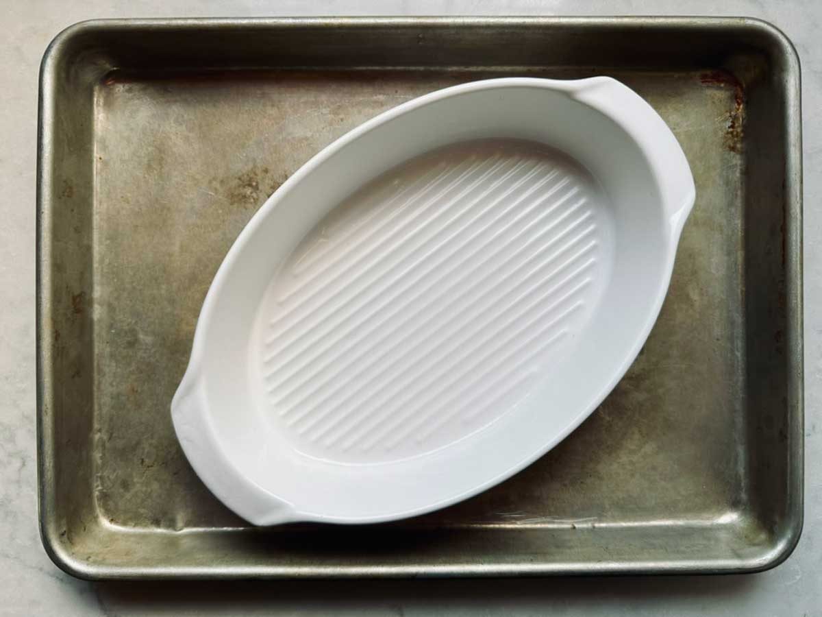 A white oval ceramic baking dish with ridges sits inside a larger rectangular metal baking sheet on a light-colored surface.
