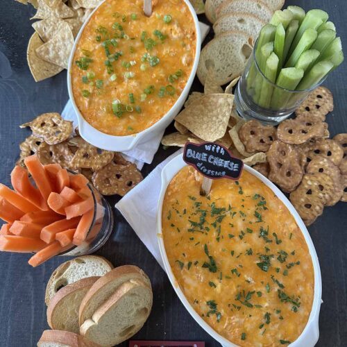 Two oval dishes of buffalo chicken dip, one with ranch and one with blue cheese, are surrounded by chips, pretzel crisps, sliced baguette, celery, and carrot sticks on a dark table. Small chalk signs label each dip.
