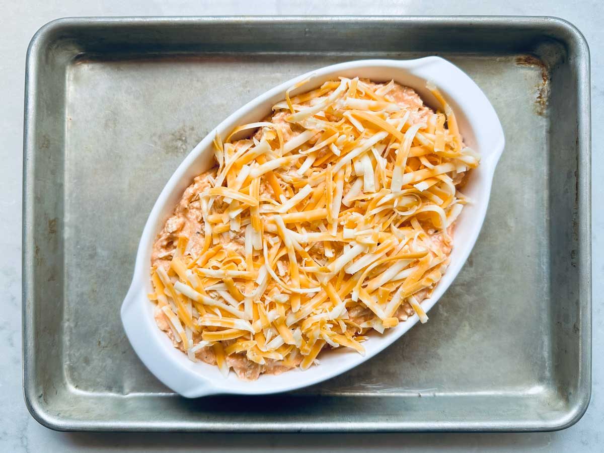 An oval white baking dish filled with a creamy mixture topped with shredded cheese, sitting on a metal baking sheet.