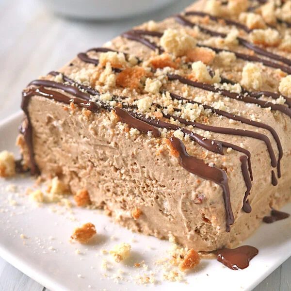 A rectangular slice of creamy, light brown ice cream dessert is topped with chocolate drizzle and crumbled cookies on a white plate, with more dessert in the background.