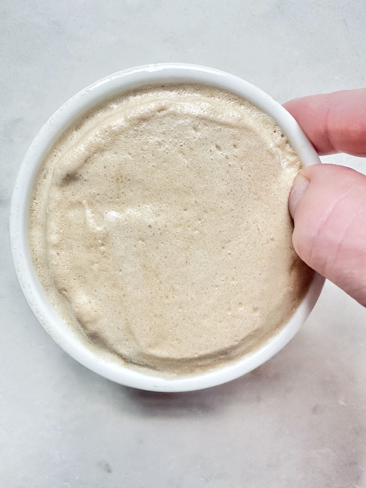 A hand holds a small white bowl filled with bubbly, creamy beige sourdough starter against a light, textured background.