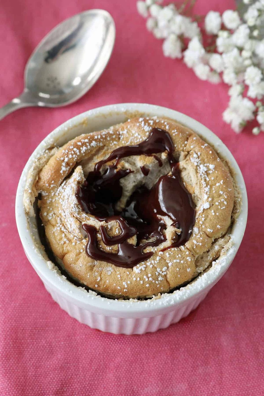 A chocolate lava cake in a white ramekin, topped with melted chocolate and powdered sugar, sits on a pink cloth next to a silver spoon and white flowers.