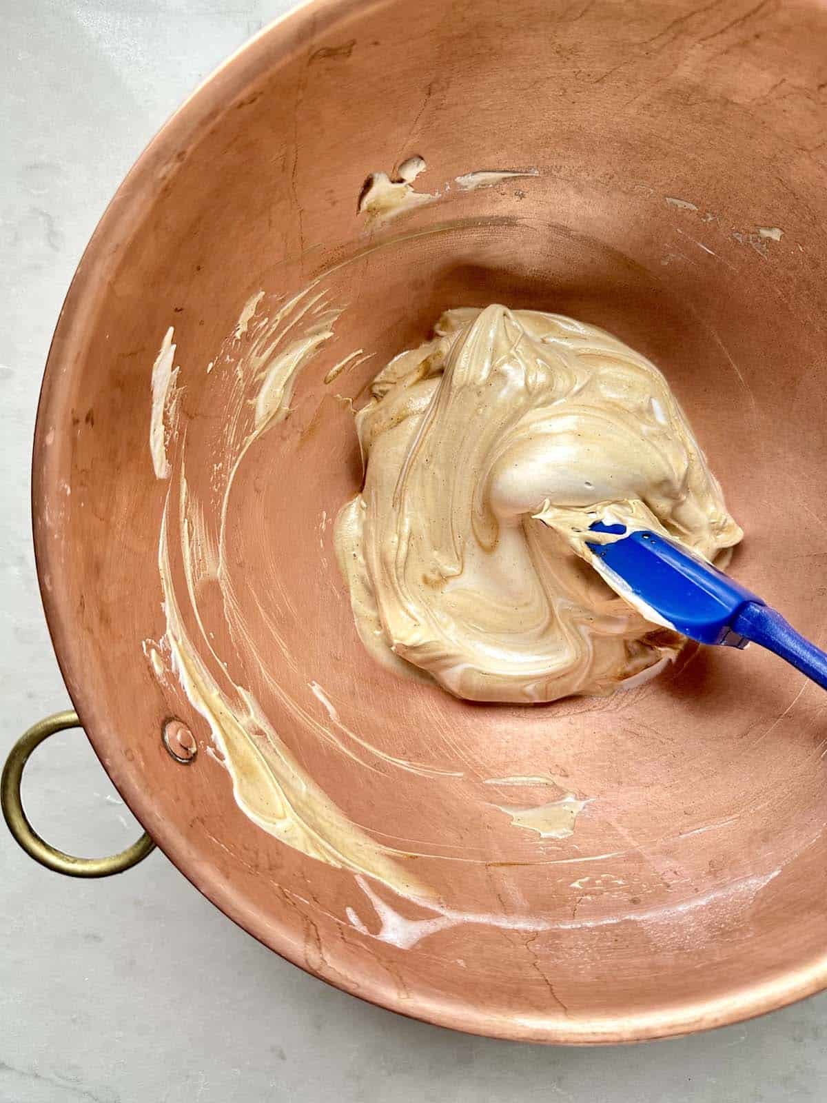A copper mixing bowl containing a thick, glossy, tan-colored mixture being stirred with a blue spatula. The sides of the bowl are partially coated with the mixture.