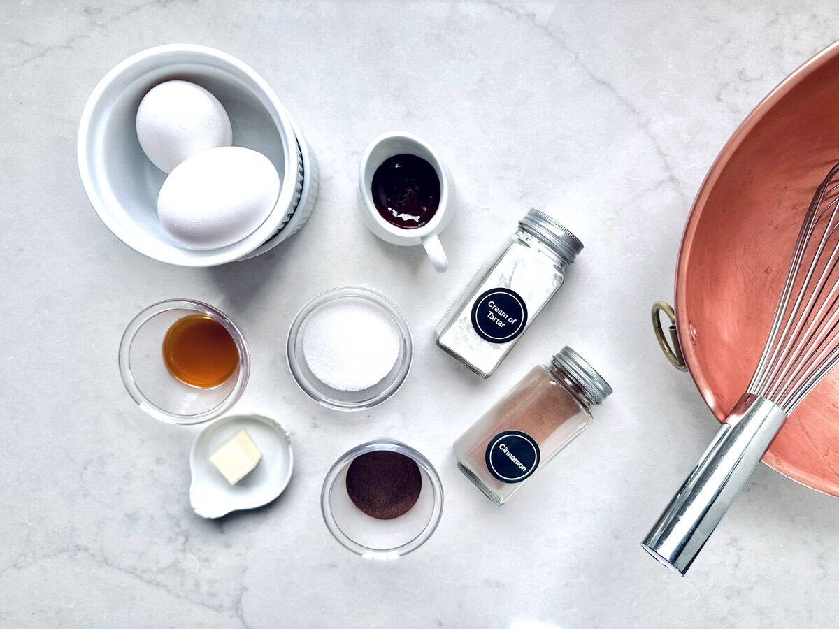 A top-down view of baking ingredients on a marble surface, including eggs, vanilla extract, sugar, butter, cocoa powder, chocolate, flour, cinnamon, and a copper mixing bowl with a whisk.