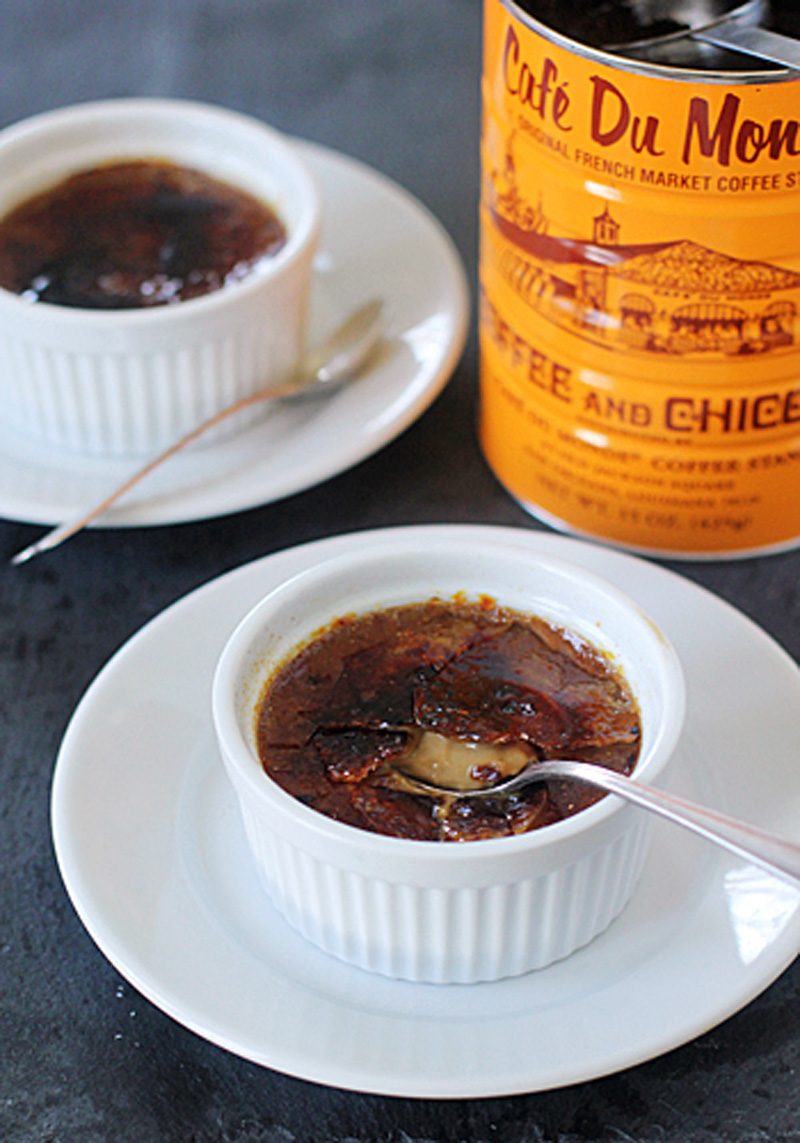 Two white ramekins of crème brûlée, each on a white plate with a spoon. One crème brûlée has a spoon breaking its caramelized sugar top. A Café Du Monde coffee can is in the background.