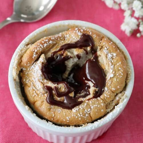 A chocolate lava cake in a white ramekin, topped with melted chocolate and powdered sugar, sits on a pink cloth next to a silver spoon and white flowers.