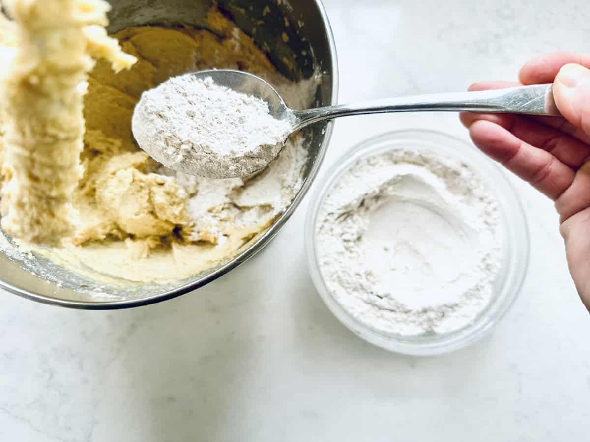 A hand holds a spoon of flour above a mixing bowl with yellow batter. A separate bowl of flour sits nearby on a white countertop.