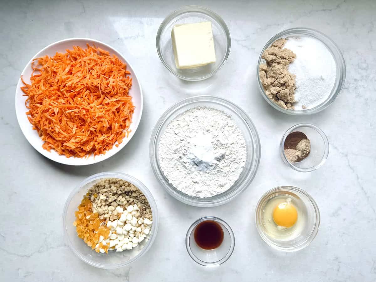 Various baking ingredients in bowls on a white surface, including shredded carrots, butter, flour with baking powder, brown and white sugars, cinnamon, nutmeg, walnuts, dried fruit, white chocolate chips, vanilla, and an egg.