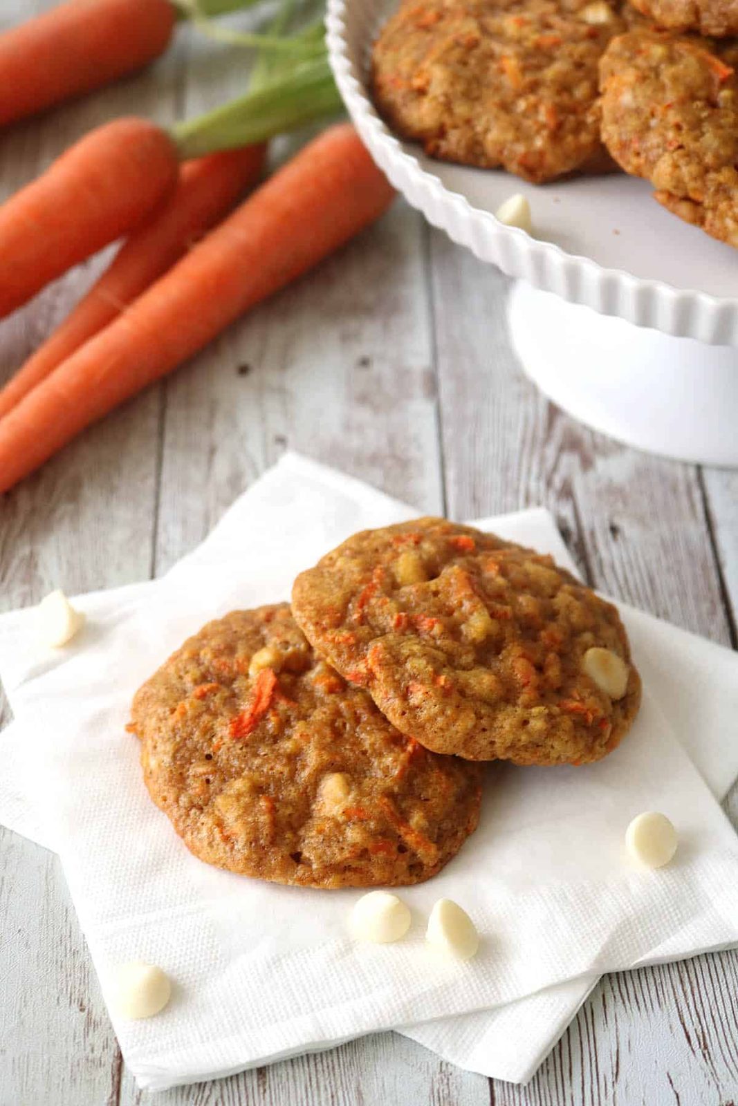 Two carrot cookies with white chocolate chips rest on a white napkin, with fresh carrots and more cookies displayed on a cake stand in the background on a rustic wooden surface.