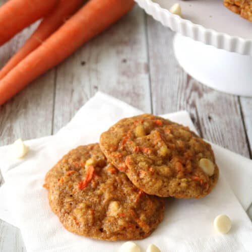 Two carrot cookies with white chocolate chips rest on a white napkin, with fresh carrots and more cookies displayed on a cake stand in the background on a rustic wooden surface.