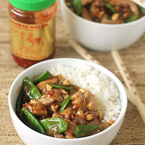 A bowl of white rice served with stir-fried chicken, snap peas, and sauce, placed next to chopsticks. Another similar bowl and a jar of chili garlic sauce are in the background on a woven surface.