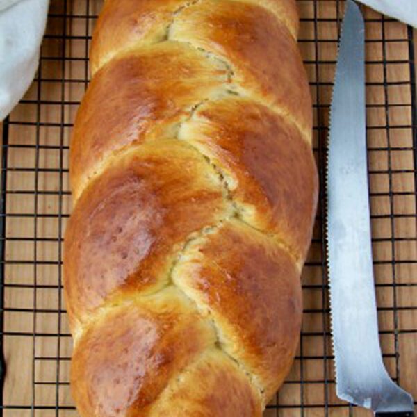A golden-brown braided loaf of bread rests on a cooling rack next to a serrated bread knife. A white cloth is partially visible at the top of the image.