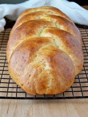 A golden-brown braided loaf of bread cooling on a black wire rack, placed on a wooden surface with a white cloth in the background.