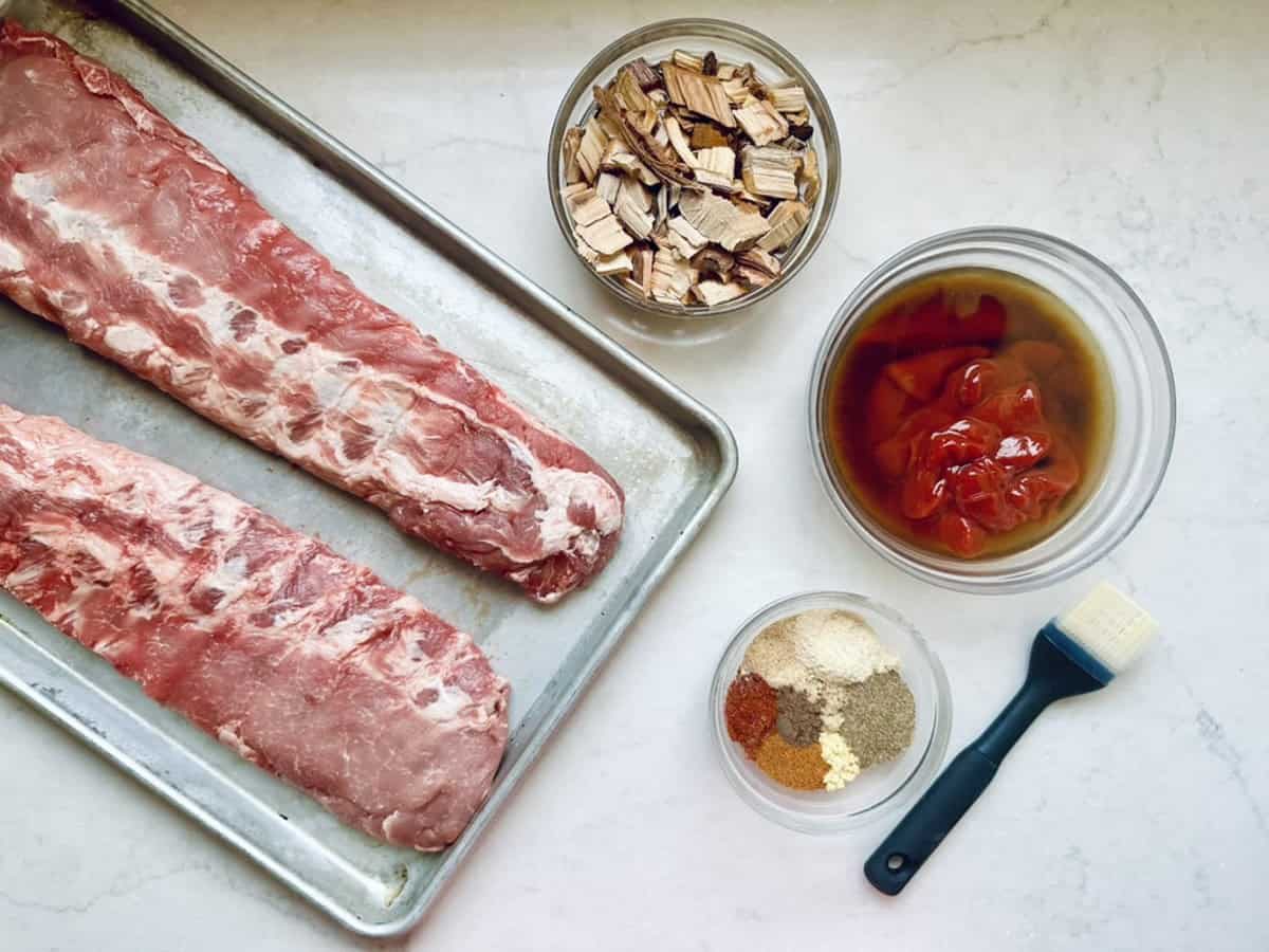 Two raw pork ribs on a metal tray next to bowls of wood chips, barbecue sauce with chopped tomatoes, assorted spices, and a basting brush on a white countertop.