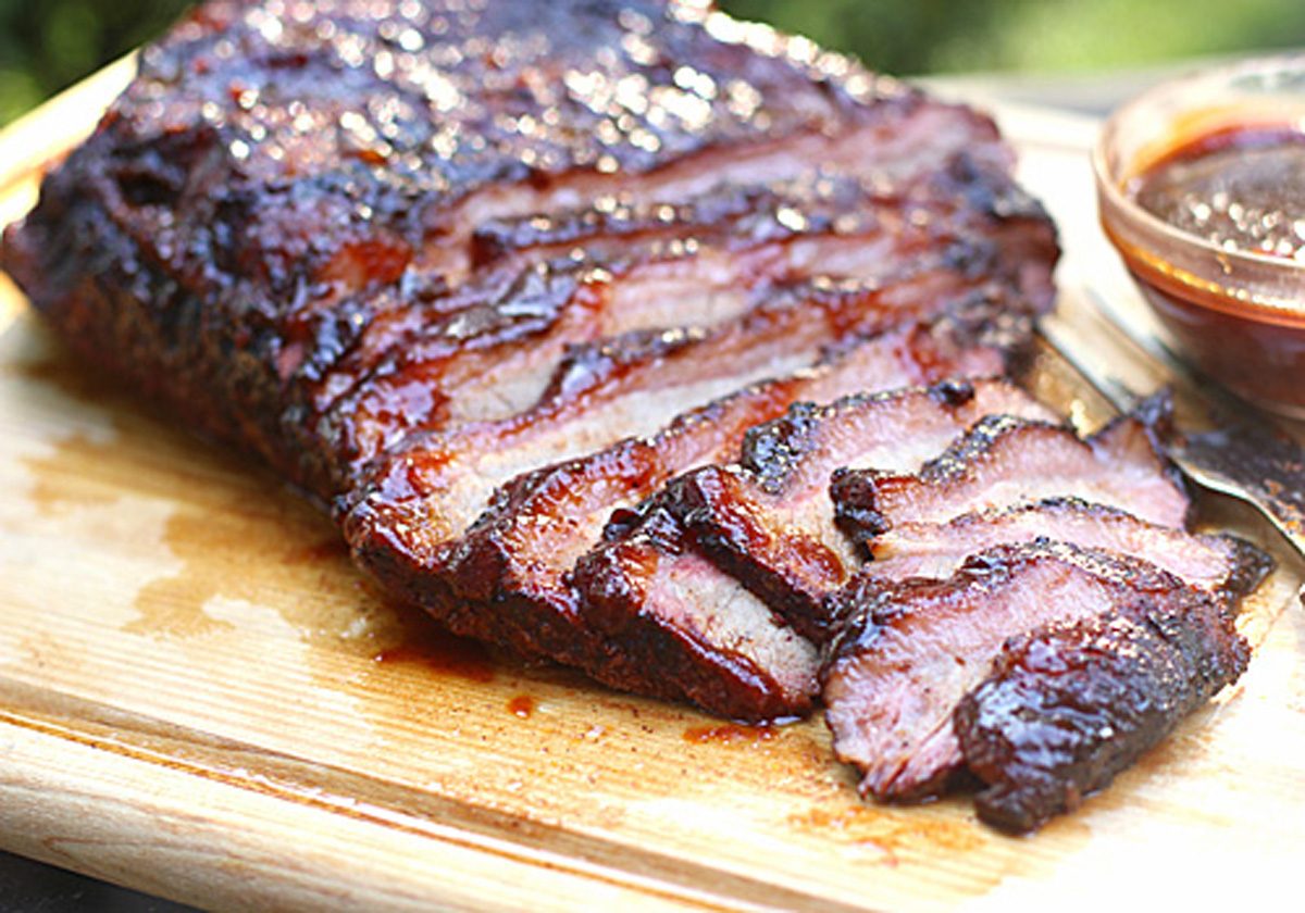 Sliced smoked brisket with a dark, caramelized crust is arranged on a wooden cutting board. A fork and a small bowl of barbecue sauce sit beside the juicy, tender meat.