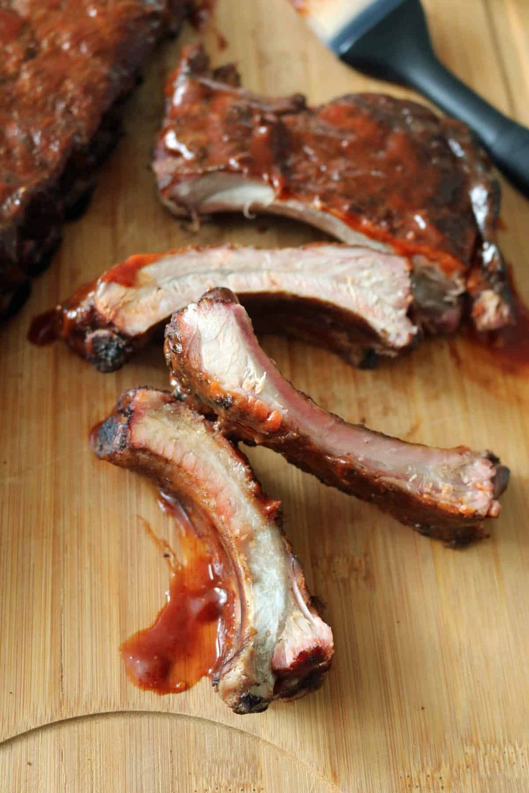 Close-up of three barbecue pork ribs with sauce on a wooden cutting board, with more ribs and a basting brush visible in the background.
