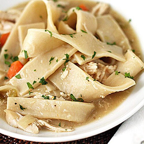 A bowl of homemade chicken and noodles with wide egg noodles, shredded chicken, carrots, and parsley in a savory broth.