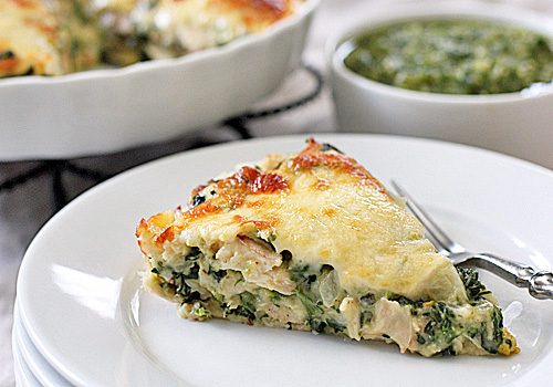 A slice of savory pie with a golden, melted cheese topping, filled with chicken and spinach, served on a white plate with a fork; a dish of green sauce is in the background.