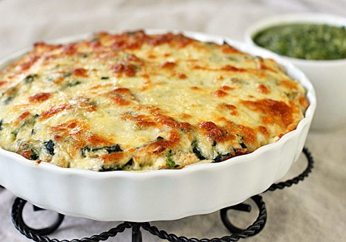 A baked cheesy casserole with a golden-brown top in a white dish, sitting on a trivet. A small bowl of green sauce is in the background.