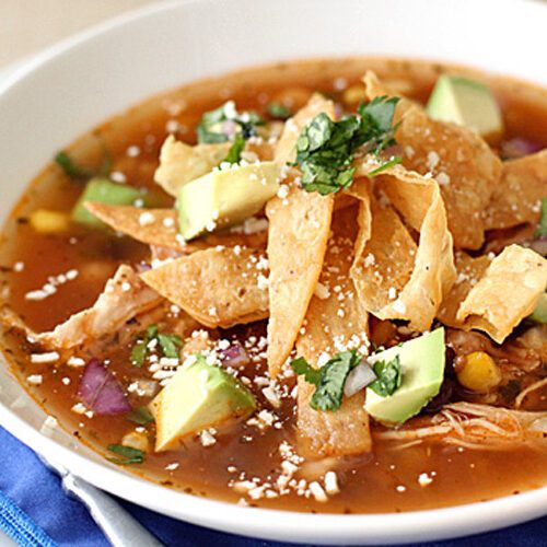 A bowl of tortilla soup topped with crispy tortilla strips, diced avocado, shredded chicken, chopped cilantro, corn, and crumbled cheese, served on a white plate with a blue napkin and spoon.