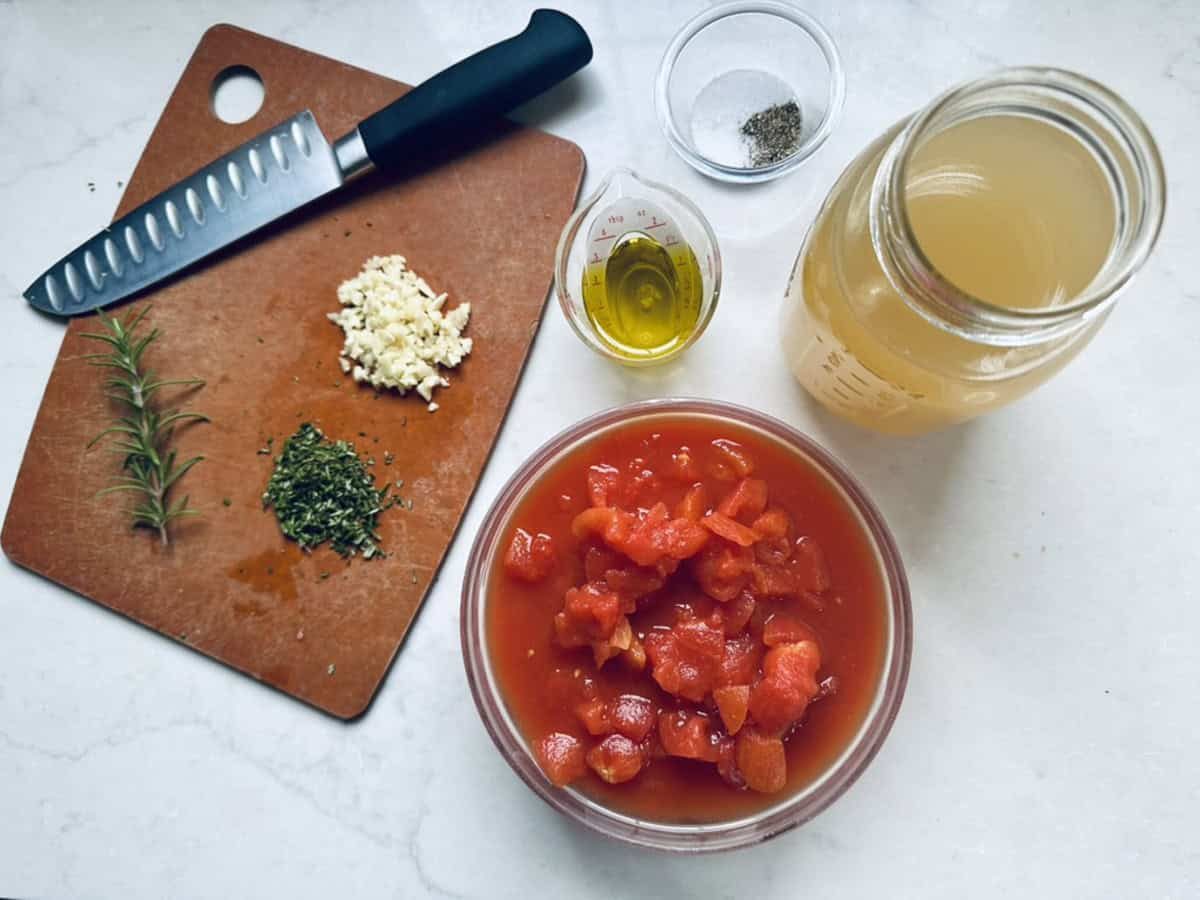 A cutting board with a knife, chopped garlic, rosemary, and dried herbs next to bowls of chopped tomatoes, olive oil, salt and pepper, and a jar of vegetable broth on a white countertop.