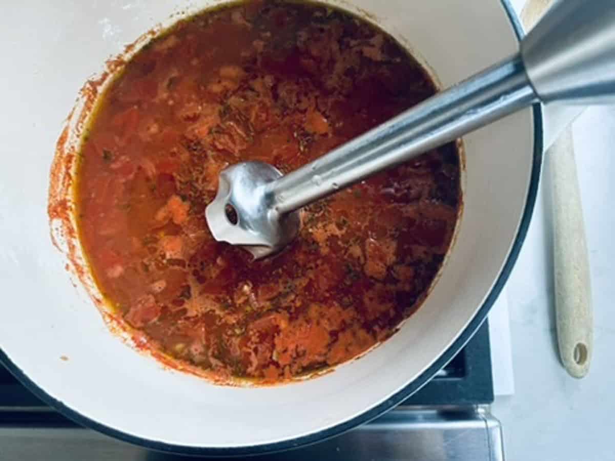 An immersion blender is blending a red tomato-based soup or sauce in a large white pot on a stove. A wooden spatula rests nearby.