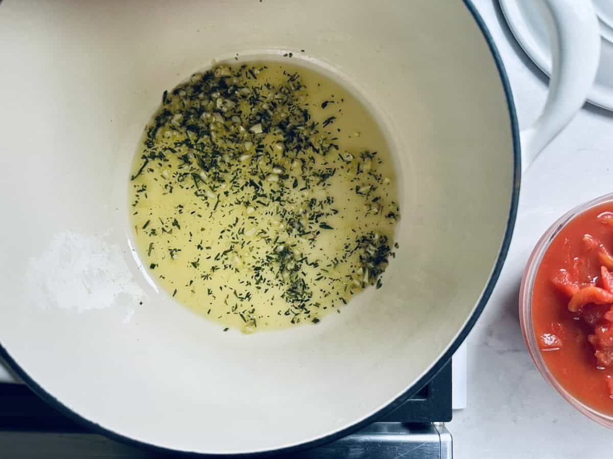 Chopped garlic and herbs sautéing in oil in a white pot on a stovetop, with a bowl of diced tomatoes next to the pot.