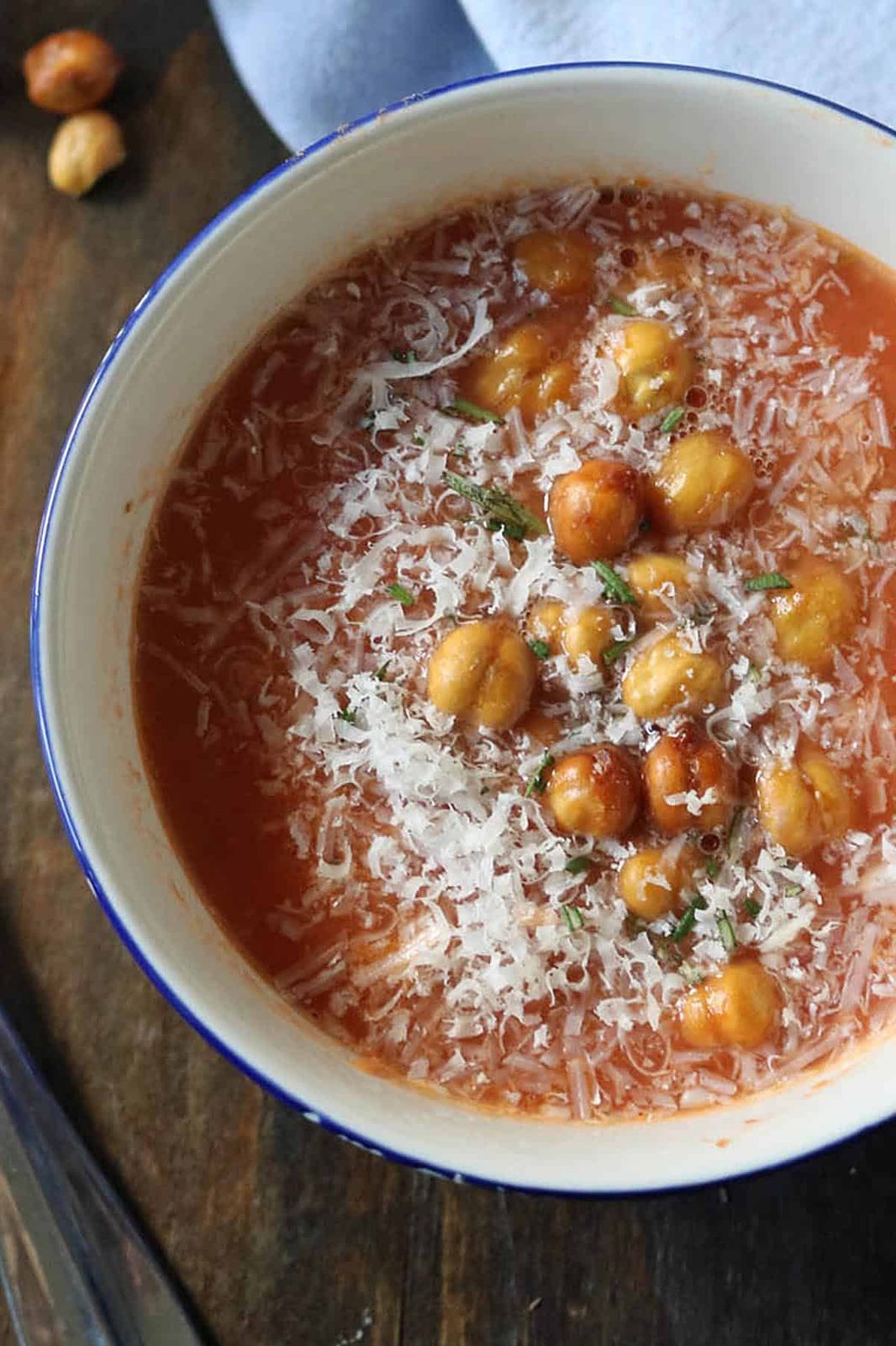 A bowl of tomato soup topped with grated cheese, roasted chickpeas, and herbs, placed on a wooden surface with a napkin partially visible in the background.