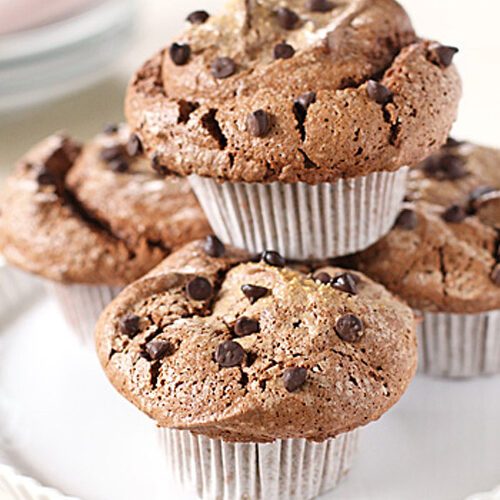 Three chocolate chip muffins in white paper liners are stacked on a white scalloped cake stand, with more muffins blurred in the background. The tops are cracked and sprinkled with mini chocolate chips.