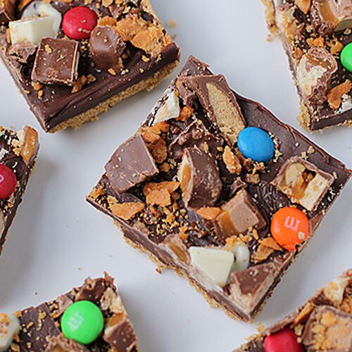Squares of chocolate dessert bars topped with assorted candy pieces, including M&Ms, nougat, and white chocolate, arranged on a white surface.