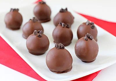 A white plate holds several round chocolate truffles, each topped with a few small chocolate chips. The plate is set on a red cloth, creating a simple yet elegant dessert presentation.