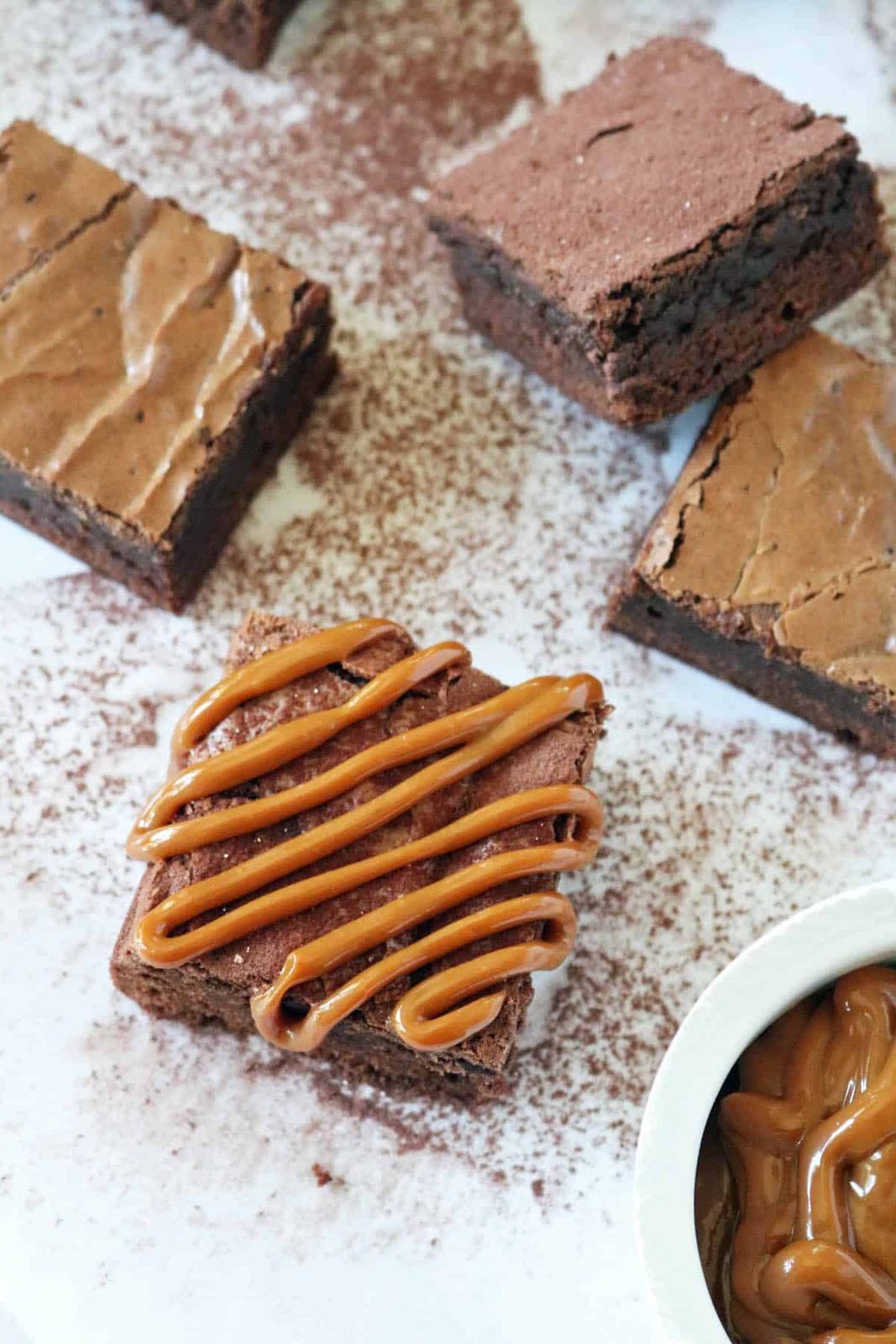 A square Chocolate Chipotle Brownie drizzled with caramel sits on a white surface dusted with cocoa powder, surrounded by more plain brownies. A small bowl of caramel is partially visible in the corner.