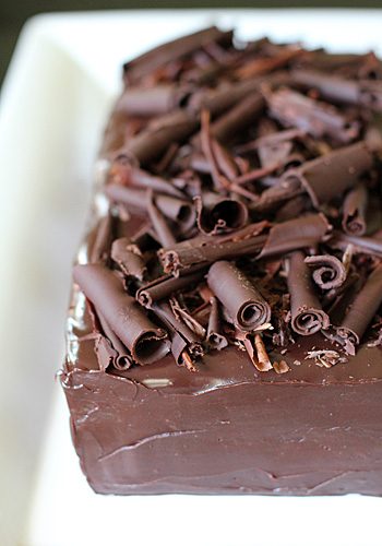 A rectangular chocolate cake covered in smooth chocolate ganache, topped with generous dark chocolate curls, sits on a white plate.