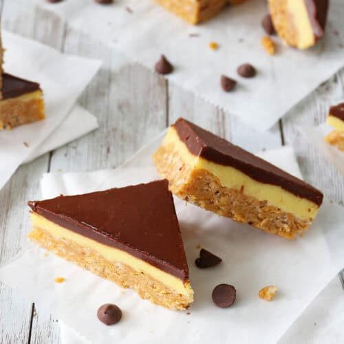 Two triangular Chocolate-Glazed Crispy Peanut Butter Pudding Bars with layers of chocolate, creamy yellow filling, and a crumbly base rest on white parchment paper. A few chocolate chips are scattered nearby on a white wooden surface.