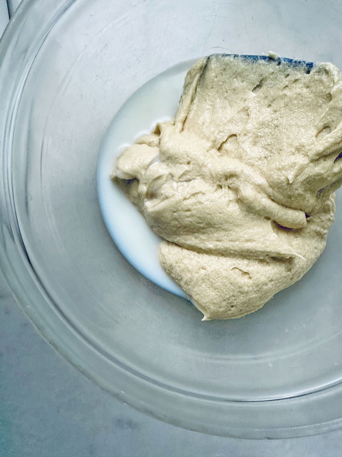 A glass bowl containing a small amount of milk with a thick, beige dough mixture being scraped into it with a spatula.