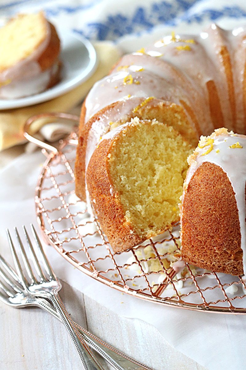 A glazed lemon bundt cake with a slice cut out, displayed on a wire rack with forks nearby. The cake is topped with icing and lemon zest, and a plate with the cut slice is in the background.