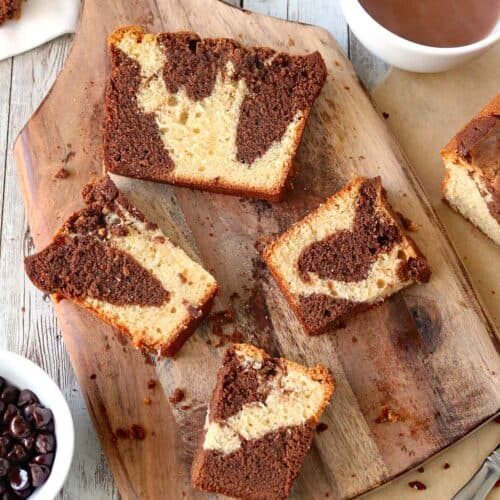 Slices of marble cake with swirls of chocolate and vanilla rest on a wooden board, surrounded by a bowl of chocolate chips and a cup of hot chocolate on a rustic white wooden table.