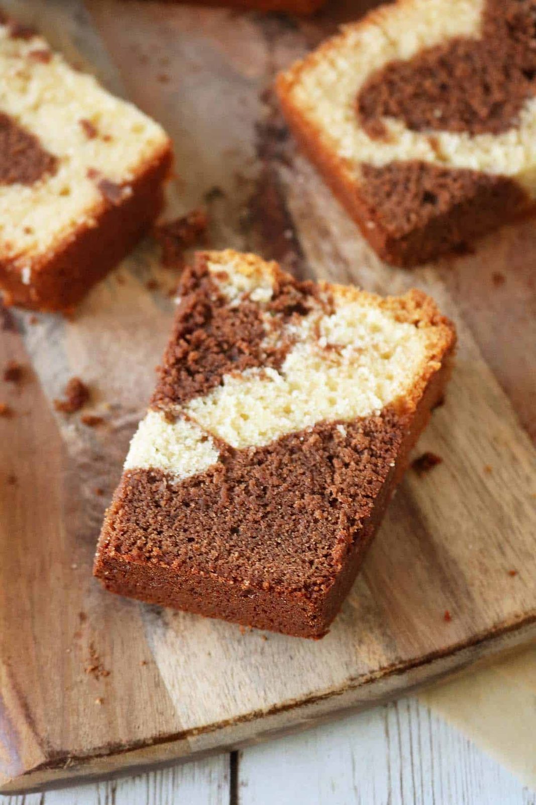 A slice of marble cake with swirls of chocolate and vanilla on a wooden board, surrounded by crumbs and other cake slices in the background.