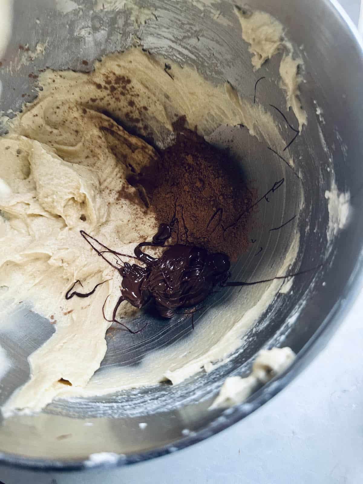 A metal mixing bowl containing creamy batter, cocoa powder, and a dollop of melted chocolate, ready to be mixed together for baking.